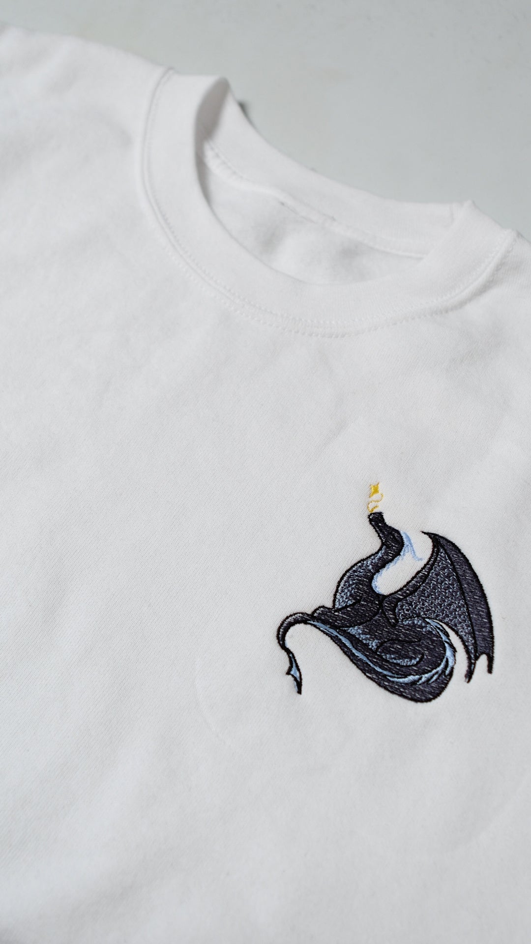 White crewneck sweatshirt with an embroidered black with blue dragon exhaling golden magical sparkles on the right chest