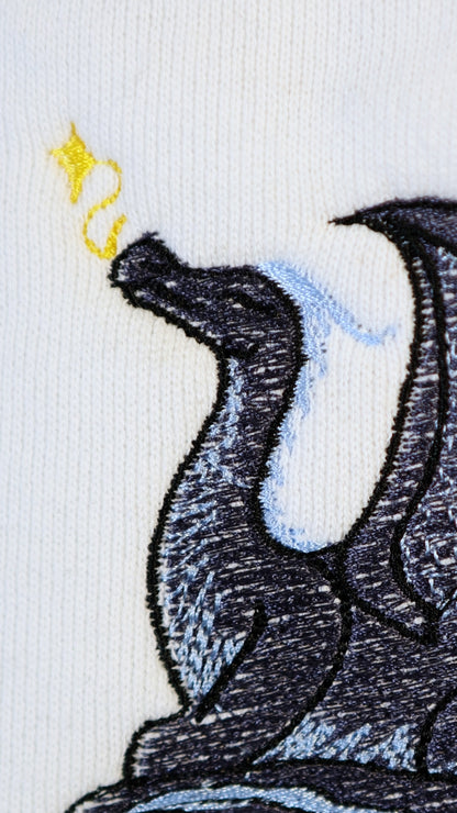 Close-up of an embroidered black and blue dragon exhaling golden magical sparkles on a white crewneck sweatshirt