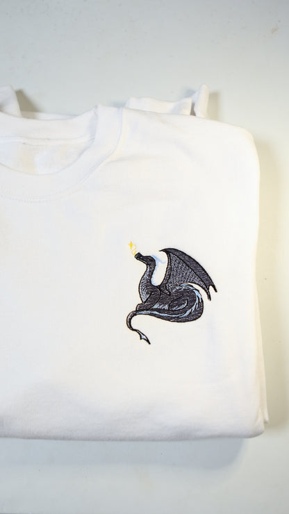 Photo of a white crewneck sweatshirt with an embroidered black and blue dragon exhaling golden magical sparkles